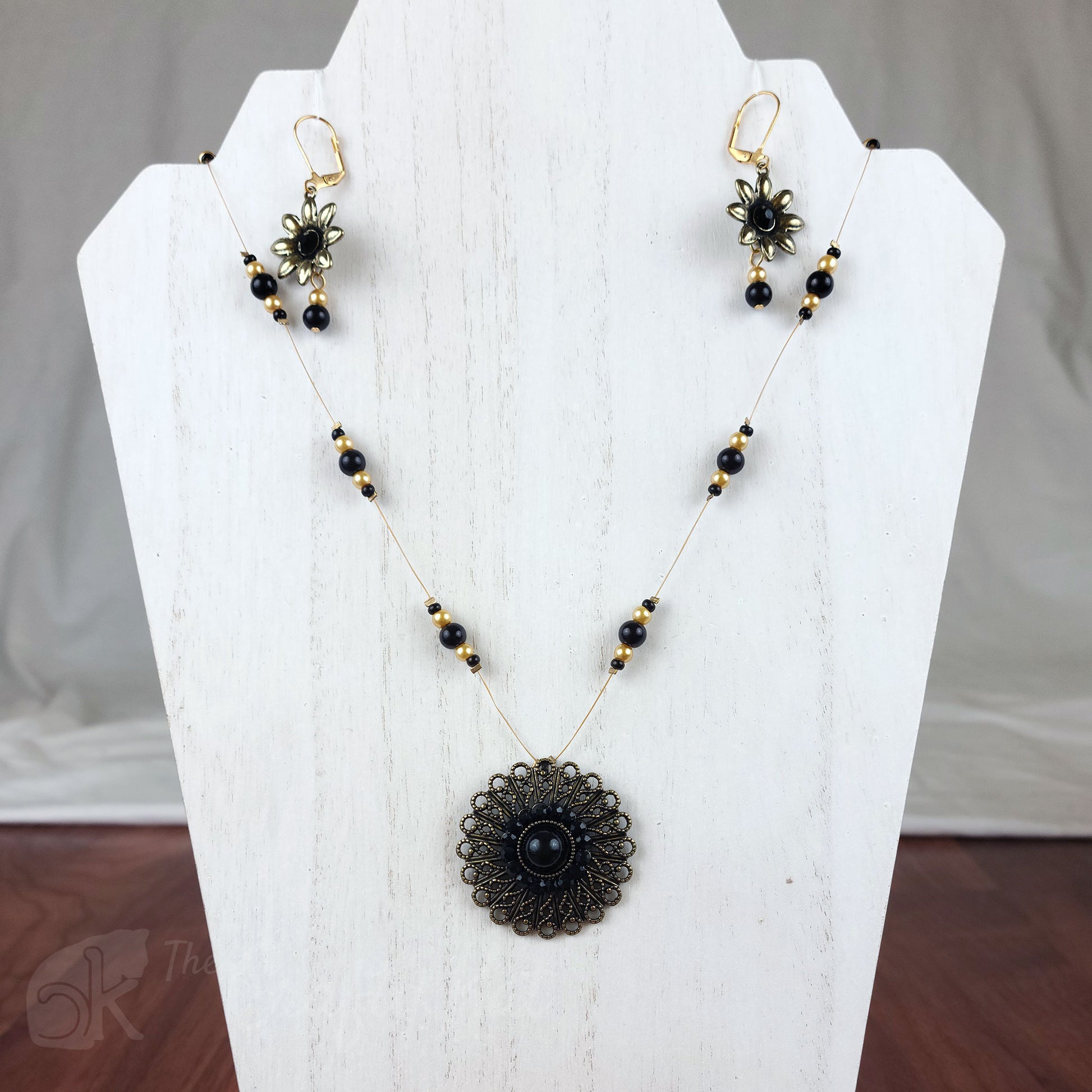 Necklace and earring set featuring antique gold flower-shaped focal pieces with black cabochons and black and gold glass pearls strung on antique gold tigertail.