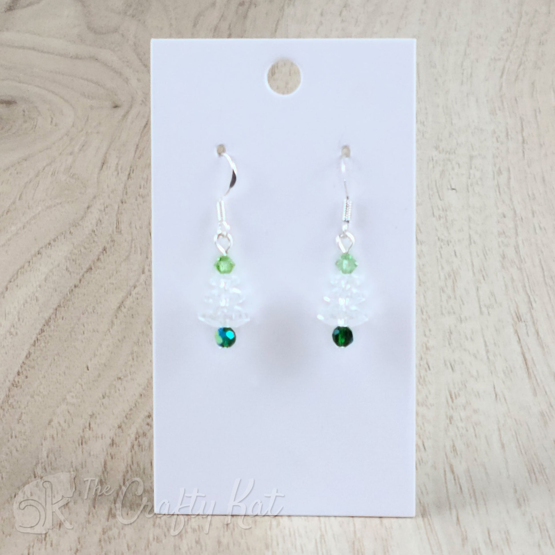 A pair of earring drops made of faceted crystal beads, each drop forming the shape of a holiday tree. This variation features an iridescent peacock green base, clear branches, and a pale green topper on silver-plated base metal.