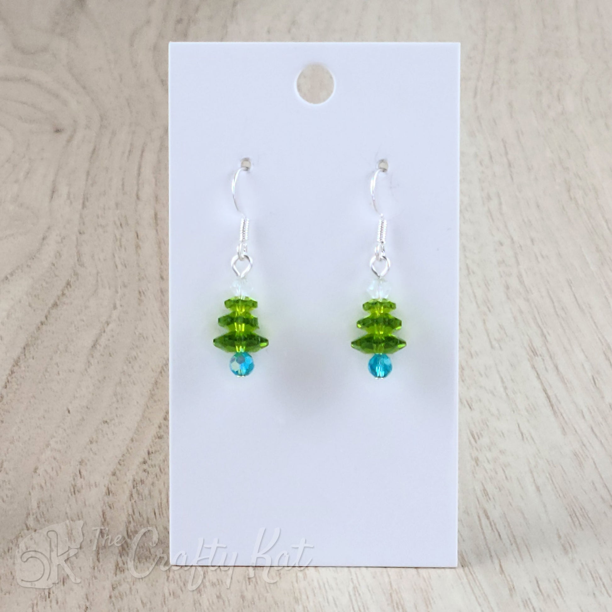 A pair of earring drops made of faceted crystal beads, each drop forming the shape of a holiday tree. This variation features an aqua/sky blue base, peridot branches, and a white topper on silver-plated base metal.