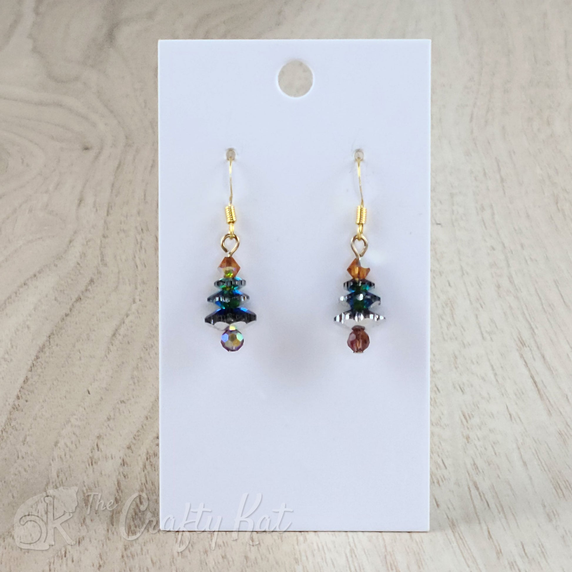A pair of earring drops made of faceted crystal beads, each drop forming the shape of a holiday tree. This variation features a deep purple base, iridescent emerald branches, and a gold topper on gold-plated base metal.