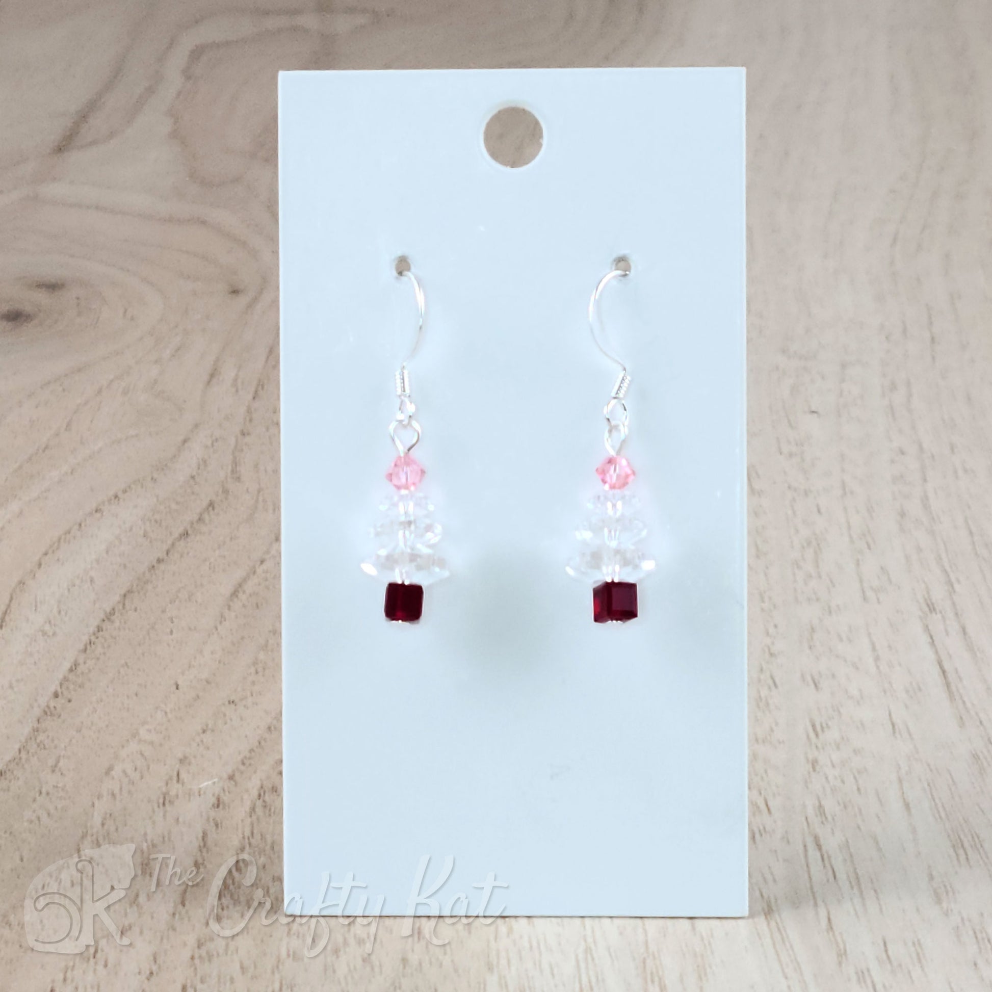 A pair of earring drops made of faceted crystal beads, each drop forming the shape of a holiday tree. This variation features a maroon base, clear branches, and a light pink topper on silver-plated base metal.