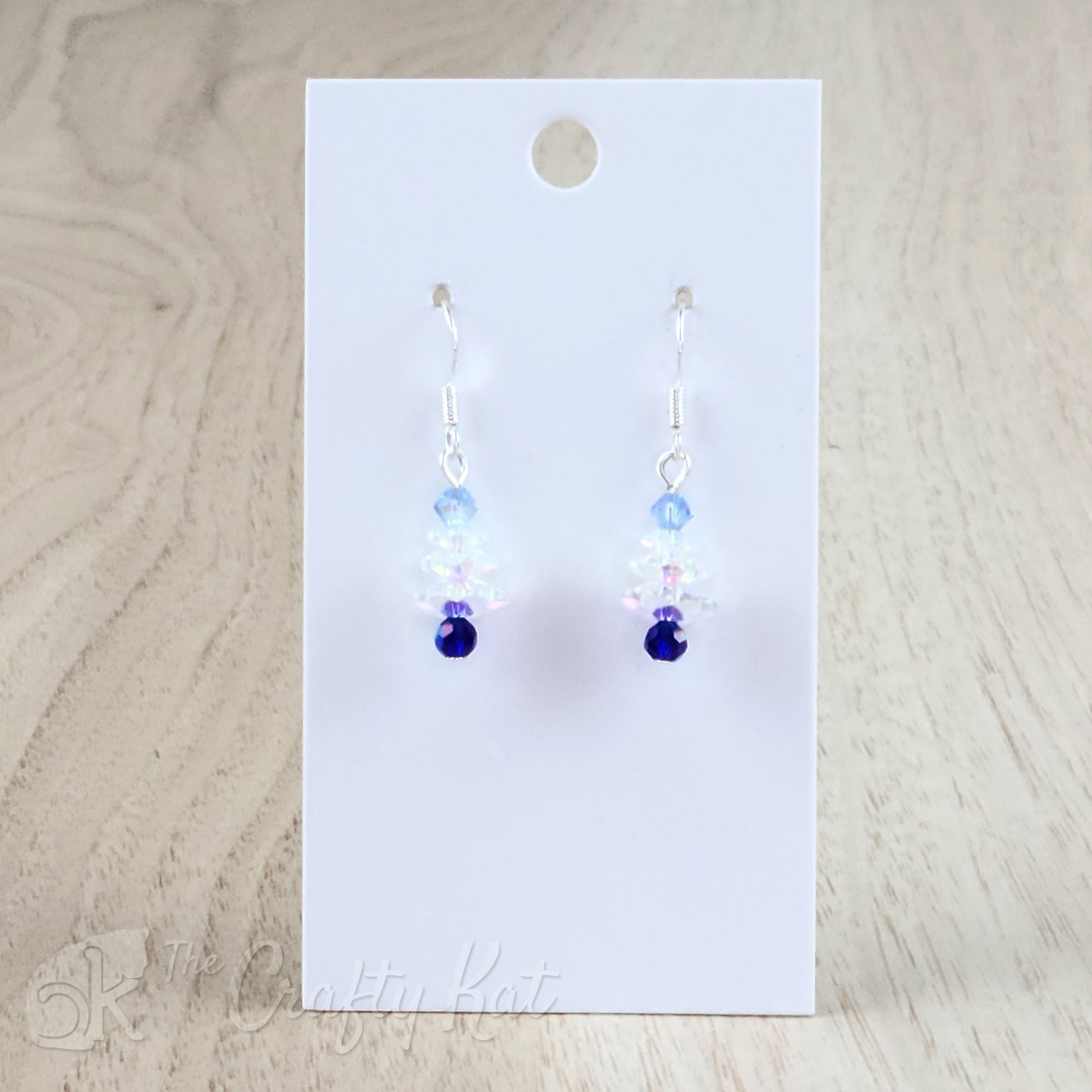 A pair of earring drops made of faceted crystal beads, each drop forming the shape of a holiday tree. This variation features an iridescent dark blue base, iridescent white branches, and a pale blue topper on silver-plated base metal.