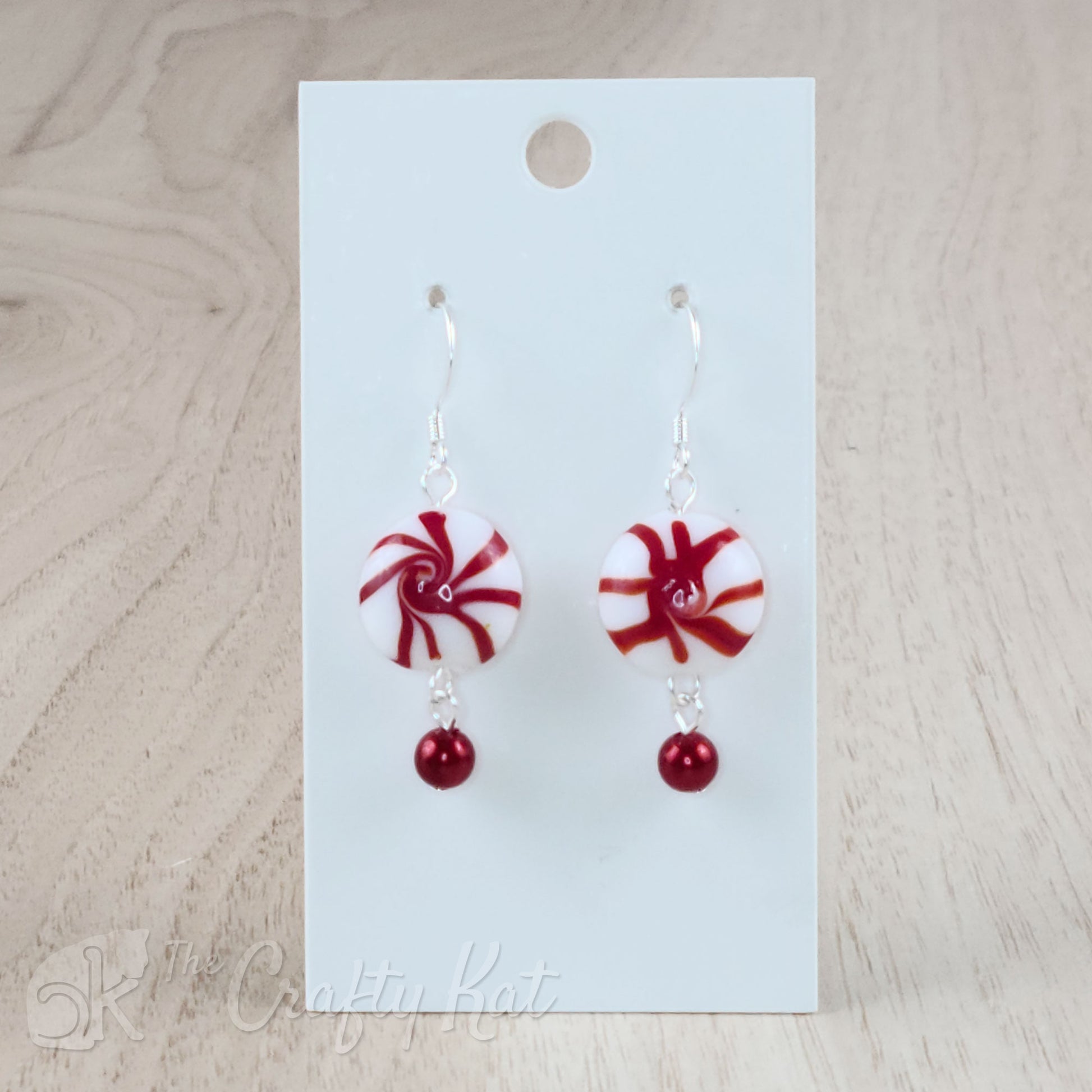 A pair of earrings featuring lampwork glass beads in the shape of classic "starlight" peppermint candies, accented with a red glass pearl on silver base metal.