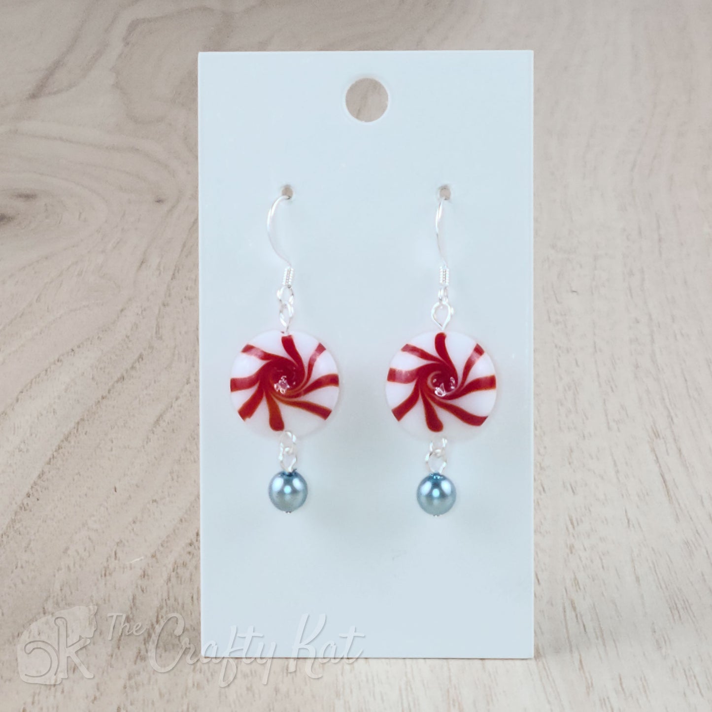 A pair of earrings featuring lampwork glass beads in the shape of classic "starlight" peppermint candies, accented with a light blue glass pearl on silver base metal.