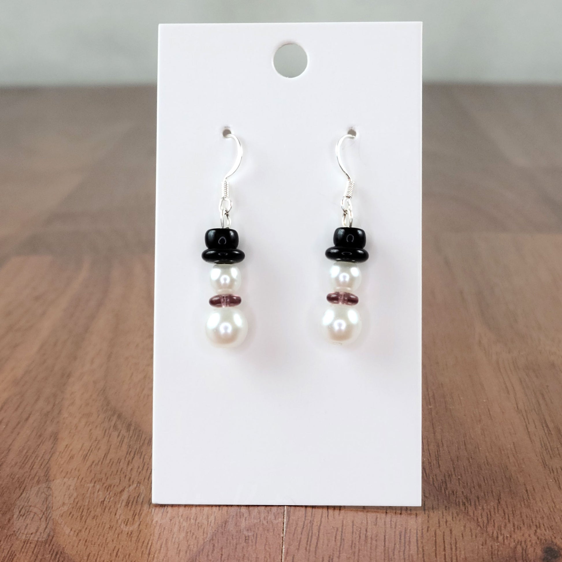 A pair of earring drops made of glass pearls and pressed glass beads, each drop forming the shape of a snowman with a purple scarf and black top hat.