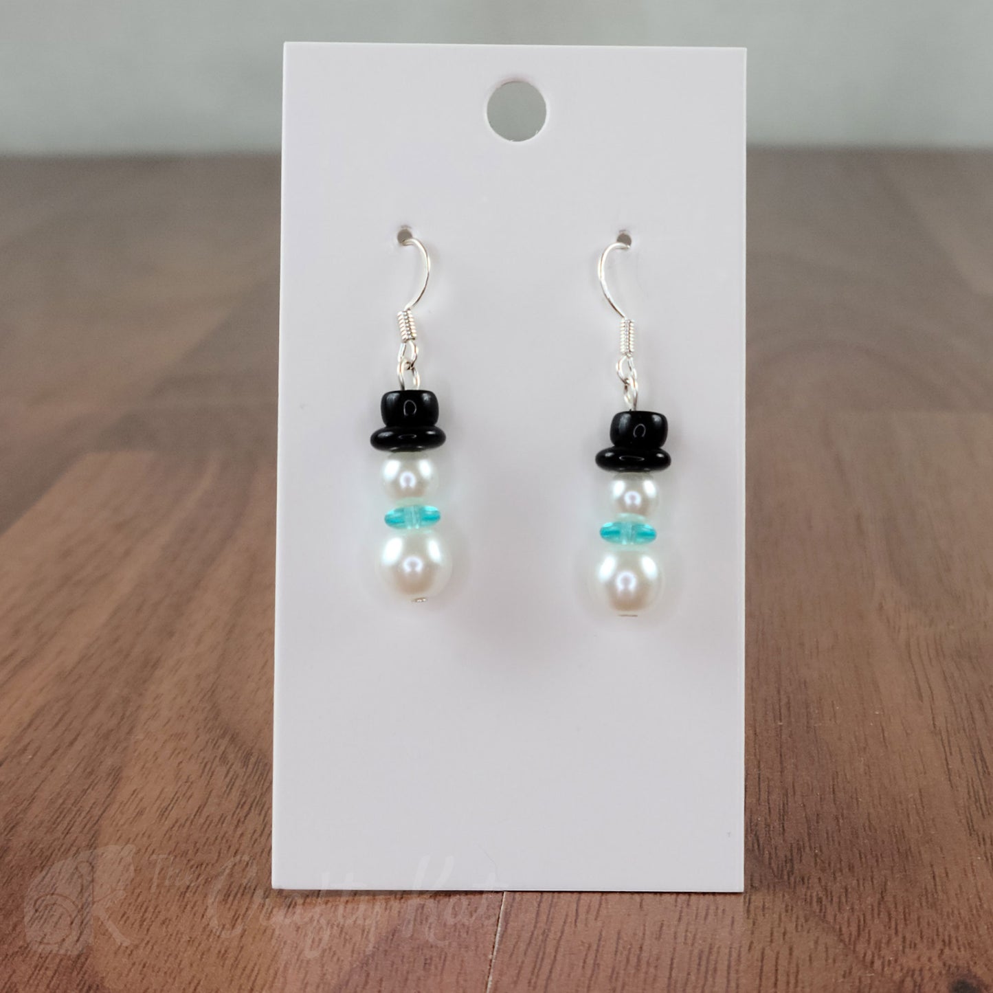 A pair of earring drops made of glass pearls and pressed glass beads, each drop forming the shape of a snowman with an aqua (sky blue) scarf and black top hat.
