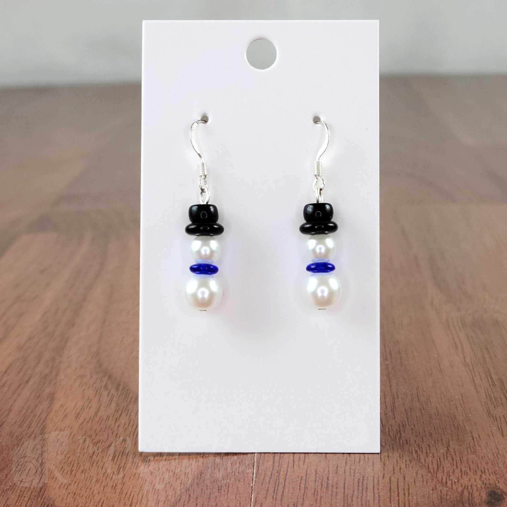 A pair of earring drops made of glass pearls and pressed glass beads, each drop forming the shape of a snowman with a blue scarf and black top hat.