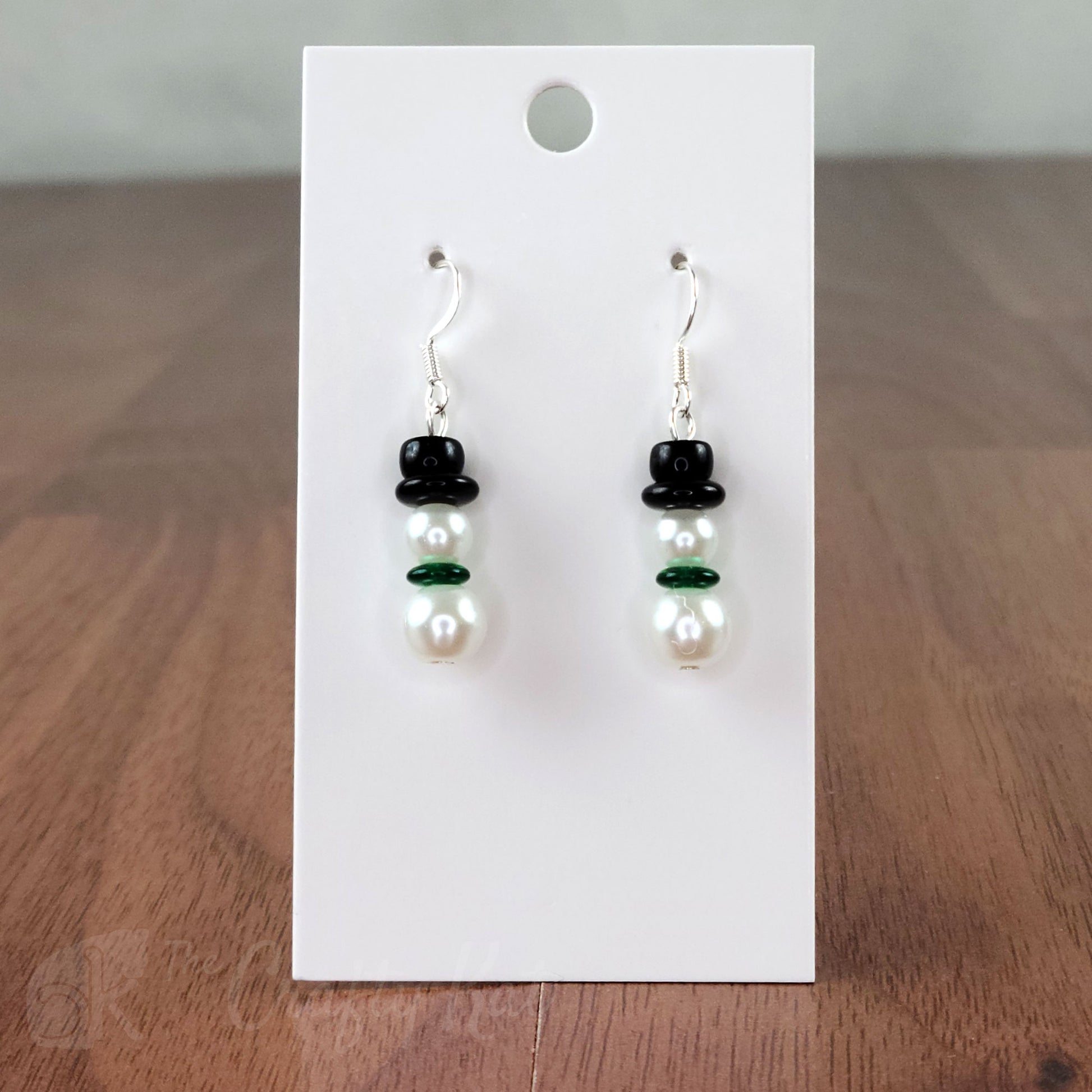 A pair of earring drops made of glass pearls and pressed glass beads, each drop forming the shape of a snowman with a green scarf and black top hat.