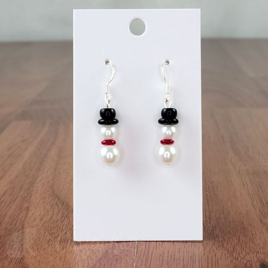 A pair of earring drops made of glass pearls and pressed glass beads, each drop forming the shape of a snowman with a red scarf and black top hat.